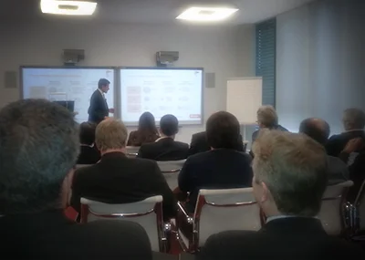 ITVT-themed scene showing a presenter speaking in front of two digital screens during a conference session, with attendees seated and listening in a modern meeting room.