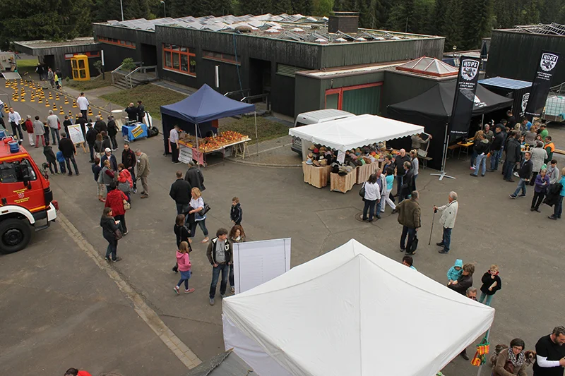 Outdoor community event in a complex with several stands under white and black canopies. People walk around and gather around tables displaying goods. In the background, dark green buildings with orange window frames and a section with yellow and black traffic cones can be seen. From ITVT.