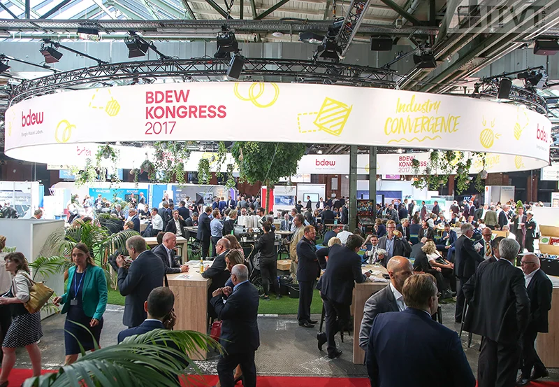 Large industry event at the BDEW Kongress 2017, showing a busy exhibition hall with numerous attendees networking, visiting booths, and engaging in discussions. The venue features a prominent circular banner overhead with event branding and modern lighting. Produced for ITVT.