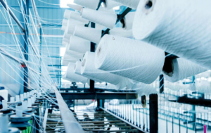 A close-up view of multiple white yarn spools arranged on a large textile machine inside a factory. Thin threads extend from the spools and are guided through the machinery, with a blurred industrial background of similar equipment and natural light filtering in