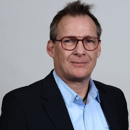 Professional portrait-style photo of a person wearing a dark suit jacket over a light blue collared shirt, used for ITVT corporate materials.
