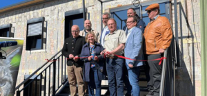 Group of people standing on outdoor stairs in front of a building during a ribbon-cutting ceremony, holding a red ribbon. The building has a rustic exterior with large windows and black railings. By ITVT.