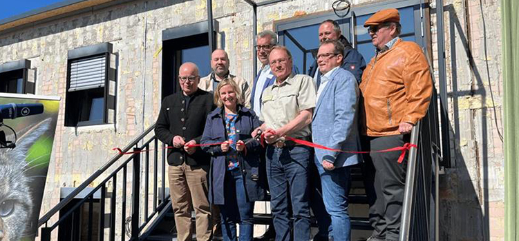 Group of people standing on outdoor stairs in front of a building during a ribbon-cutting ceremony, holding a red ribbon. The building has a rustic exterior with large windows and black railings. By ITVT.