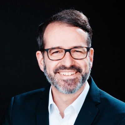A professional studio portrait of a person wearing a dark suit jacket and a white collared shirt, photographed against a black background. Image prepared for ITVT corporate branding and materials.