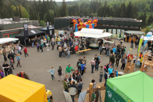 GroÃŸes Outdoor-Community-Event in einer von BÃ¤umen umgebenen Anlage mit mehreren StÃ¤nden unter weiÃŸen, schwarzen und grÃ¼nen Ãœberdachungen. Im Hintergrund ist eine farbenfrohe aufblasbare Struktur zu sehen, zusammen mit Informationsdisplays und einem Imbissstand. Ãœberall auf dem GelÃ¤nde haben sich Menschen versammelt, die sich fÃ¼r die Ausstellungen und AktivitÃ¤ten interessieren. Von ITVT.