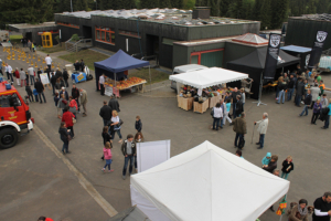 Outdoor-Community-Veranstaltung in einem Komplex mit mehreren StÃ¤nden unter weiÃŸen und schwarzen Ãœberdachungen. Die Menschen laufen herum und versammeln sich um Tische, auf denen Waren ausgestellt sind. Im Hintergrund sind dunkelgrÃ¼ne GebÃ¤ude mit orangefarbenen Fensterrahmen und ein Bereich mit gelben und schwarzen Verkehrskegeln zu sehen. Von ITVT.