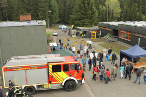 Veranstaltung im Freien in einer von BÃ¤umen umgebenen Anlage, im Vordergrund ein roter und gelber Feuerwehrwagen. Die Menschen versammeln sich um verschiedene StÃ¤nde und AusstellungsstÃ¼cke, darunter ein blaues Zelt und ein Bereich mit gelben und schwarzen Verkehrskegeln. Von ITVT.