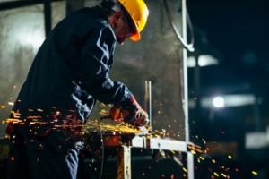 Arbeiter mit Schutzhelm, der mit einem Elektrowerkzeug Metall schleift und dabei helle Funken in einer industriellen Umgebung erzeugt.