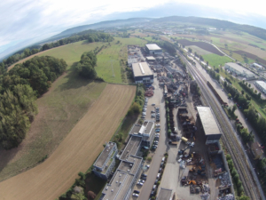Luftaufnahme eines Industriegebiets mit LagerhÃ¤usern, geparkten Fahrzeugen und Eisenbahnschienen neben grÃ¼nen Feldern und HÃ¼geln im Hintergrund.