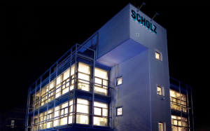 Modernes GebÃ¤ude bei Nacht mit beleuchteten Fenstern und dem Namen â€žSCHOLZâ€œ oben.