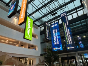 GroÃŸes Innenatrium mit hÃ¤ngenden vertikalen Bannern, auf denen â€žMicrosoftâ€œ-Logos und eine AnkÃ¼ndigung der ITVT-Roadshow fÃ¼r MÃ¼nchen vom 12.11.2024 zu sehen sind, die fÃ¼r KI im Vertrieb und Kundenservice wirbt.