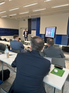 GeschÃ¤ftsprÃ¤sentation in einem modernen Konferenzraum mit Teilnehmern, die an Tischen sitzen, ihre Laptops geÃ¶ffnet haben und einem Redner, der Folien auf einer Leinwand prÃ¤sentiert.