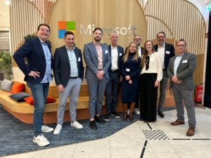 Das Foto zeigt acht Personen, die in einer Reihe in einem Raum stehen, der wie ein BÃ¼ro oder Veranstaltungsraum von Microsoft aussieht. Hinter ihnen befindet sich ein groÃŸes â€žMicrosoftâ€-Logo auf einer Holzwand mit einem bunten Microsoft-Fenstersymbol Ã¼ber dem Namen. Die Gruppe besteht aus fÃ¼nf MÃ¤nnern und drei Frauen, die alle Business-Casual-Kleidung tragen.