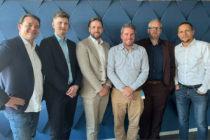 Eine Gruppe von sechs MÃ¤nnern, die gemeinsam vor einer blauen strukturierten Wand posieren und Business-Casual-Kleidung tragen.