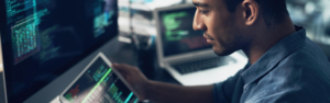Eine Person, die an einem Arbeitsplatz mit mehreren Monitoren arbeitet, auf denen Codezeilen und digitale Dashboards angezeigt werden, und ein Tablet mit Systemdaten in der Hand hÃ¤lt. Die Szene steht fÃ¼r den Fokus von ITVT auf CRM, Technologie und digitale LÃ¶sungen.