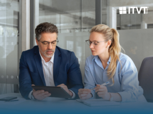 Zwei GeschÃ¤ftsleute, ein Mann in einem blauen Anzug und eine Frau in einem hellblauen Hemd, beide mit Brille, Ã¼berprÃ¼fen Informationen auf einem Tablet in einer BÃ¼roumgebung. Das Logo der ITVT Group ist in der oberen rechten Ecke zu sehen.