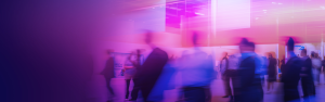 ITVT-Themenbild, das Menschen zeigt, die sich durch einen modernen Veranstaltungs- oder Ausstellungsraum bewegen, aufgenommen mit dynamischer Bewegung und beleuchtet von violettem, rosa und blauem Licht.