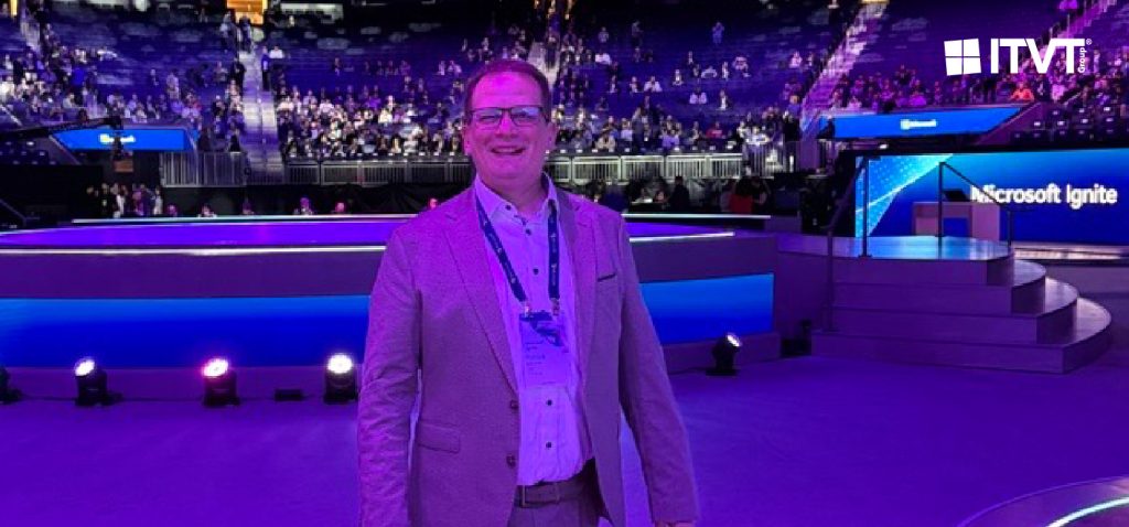 Eine Person in einem Anzug steht wÃ¤hrend der Microsoft Ignite auf der HauptbÃ¼hne einer groÃŸen, violett beleuchteten Arena. Im Hintergrund sind die BÃ¼hne, die Beleuchtung und das sitzende Publikum zu sehen, wobei ITVT auf der Veranstaltung vertreten ist.