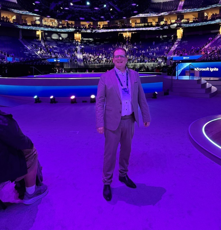 Eine Person in einem Anzug steht wÃ¤hrend der Microsoft Ignite auf der HauptbÃ¼hne einer groÃŸen, violett beleuchteten Arena. Im Hintergrund sind die BÃ¼hne, die Beleuchtung und das sitzende Publikum zu sehen, wobei ITVT auf der Veranstaltung vertreten ist.