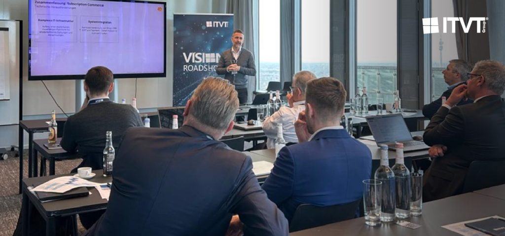 Ein professioneller Workshop oder eine PrÃ¤sentation in einem modernen Konferenzraum. Mehrere Teilnehmer sitzen an Tischen und blicken auf einen Referenten, der neben einem groÃŸen Bildschirm steht, auf dem eine Folie gezeigt wird. Neben dem Bildschirm befindet sich ein vertikales ITVT-Banner mit dem Titel â€žVISION ROADSHOWâ€. Auf den Tischen stehen Wasserflaschen, NotizbÃ¼cher, Laptops und gedruckte Unterlagen. Durch die groÃŸen, vom Boden bis zur Decke reichenden Fenster im Hintergrund hat man einen Blick auf die Stadt.