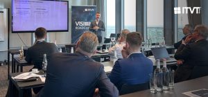 Ein professioneller Workshop oder eine PrÃ¤sentation in einem modernen Konferenzraum. Mehrere Teilnehmer sitzen an Tischen und blicken auf einen Referenten, der neben einem groÃŸen Bildschirm steht, auf dem eine Folie gezeigt wird. Neben dem Bildschirm befindet sich ein vertikales ITVT-Banner mit dem Titel â€žVISION ROADSHOWâ€. Auf den Tischen stehen Wasserflaschen, NotizbÃ¼cher, Laptops und gedruckte Unterlagen. Durch die groÃŸen, vom Boden bis zur Decke reichenden Fenster im Hintergrund hat man einen Blick auf die Stadt.