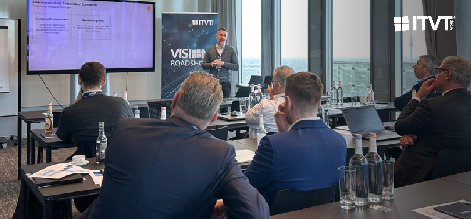 Ein professioneller Workshop oder eine PrÃ¤sentation in einem modernen Konferenzraum. Mehrere Teilnehmer sitzen an Tischen und blicken auf einen Referenten, der neben einem groÃŸen Bildschirm steht, auf dem eine Folie gezeigt wird. Neben dem Bildschirm befindet sich ein vertikales ITVT-Banner mit dem Titel â€žVISION ROADSHOWâ€. Auf den Tischen stehen Wasserflaschen, NotizbÃ¼cher, Laptops und gedruckte Unterlagen. Durch die groÃŸen, vom Boden bis zur Decke reichenden Fenster im Hintergrund hat man einen Blick auf die Stadt.
