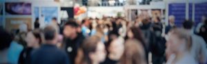 Ein Foto einer belebten Indoor-Veranstaltung oder Ausstellungshalle, in der viele Menschen herumlaufen und in Gruppen stehen. Im Hintergrund sind helle Lichter, StÃ¤nde und AusstellungsstÃ¼cke zu sehen, die eine lebhafte und geschÃ¤ftige AtmosphÃ¤re schaffen.