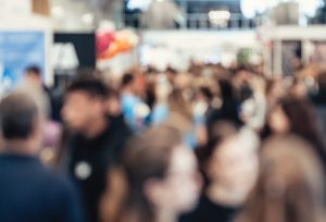 Ein Foto einer belebten Indoor-Veranstaltung oder Ausstellungshalle, in der viele Menschen herumlaufen und in Gruppen stehen. Im Hintergrund sind helle Lichter, StÃ¤nde und AusstellungsstÃ¼cke zu sehen, die eine lebhafte und geschÃ¤ftige AtmosphÃ¤re schaffen.