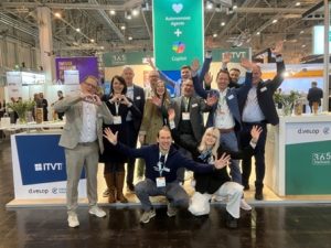 Eine Gruppe von Menschen posiert voller Energie fÃ¼r ein Gruppenfoto am Messestand von ITVT. Der Stand ist mit auffÃ¤lligen Schildern ausgestattet, darunter Banner fÃ¼r ITVT, d.velop und 365 Stadtwerk. Ein groÃŸes grÃ¼nes Display im Hintergrund wirbt fÃ¼r â€žAutonomous Agents + Copilotâ€. Die Szene spielt in einer belebten Messehalle, in der Ã¼berall Lichter, StÃ¤nde und Besucher zu sehen sind.